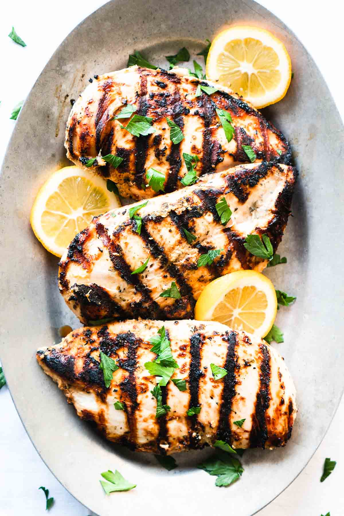 An oval platter with grilled Greek yogurt marinated chicken breasts, lemon slices and fresh herbs.