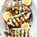 A plate with grilled Greek yogurt marinated chicken breasts, lemon slices and fresh herbs.