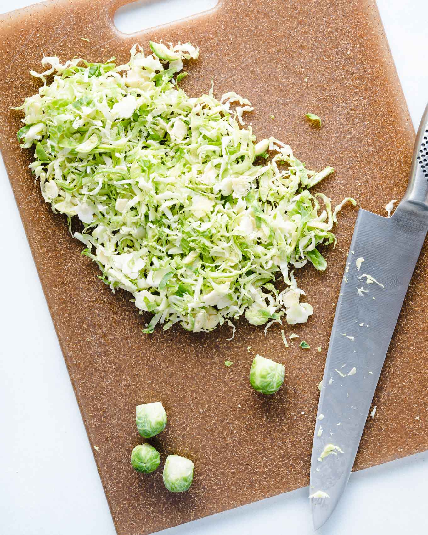 raw brussels sprouts being chopped by a sharp knife on a brown cutting board