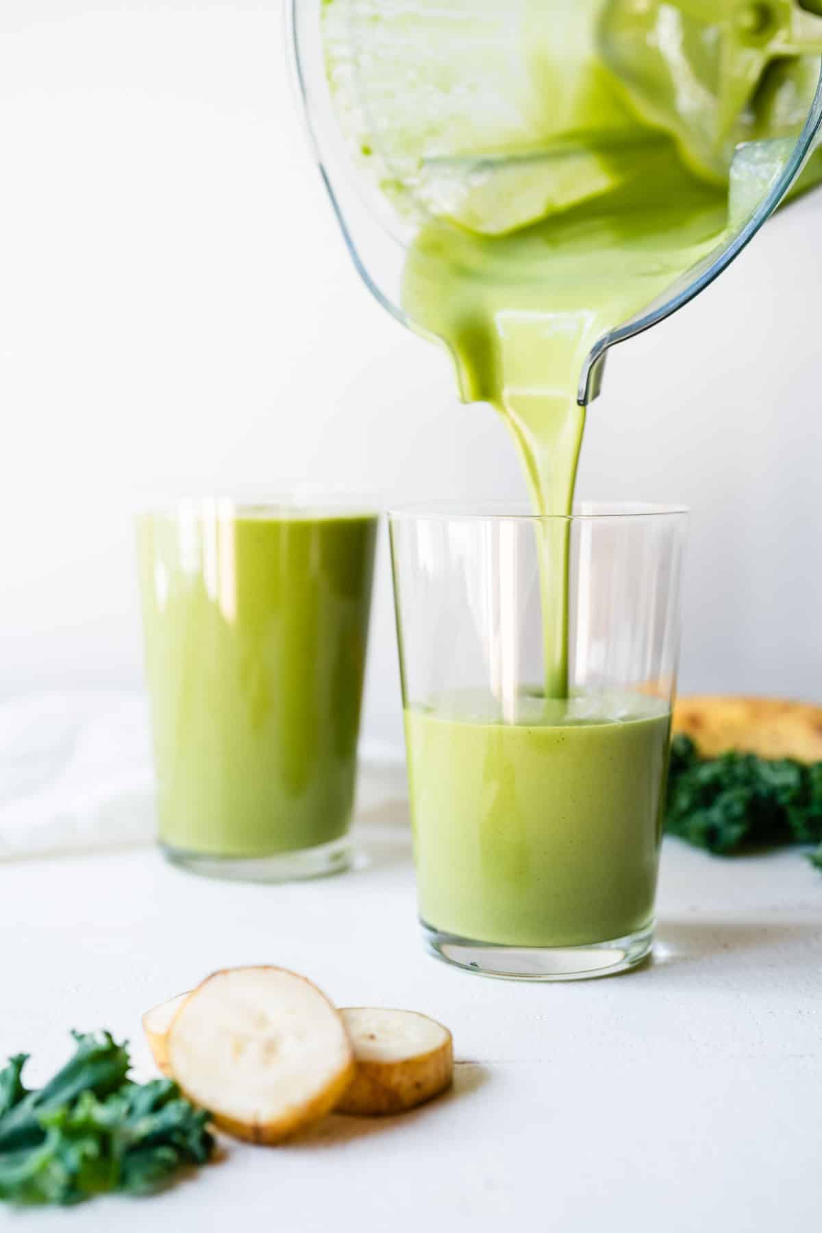 A blender cup pouring a banana kale smoothie into two glass cups.