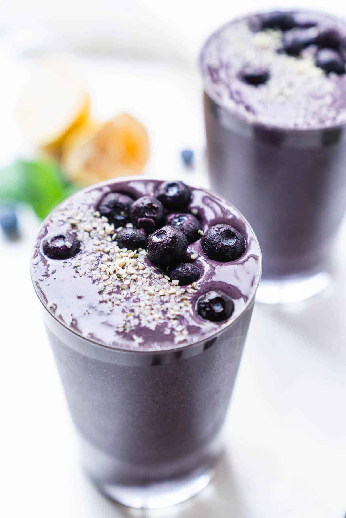 Two smoothies in clear glasses on a white table with fresh fruit on the side.