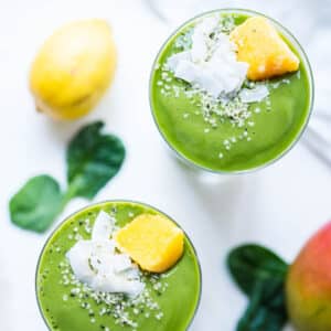 Two mango spinach smoothies on a white table, topped with fresh fruit and coconut.