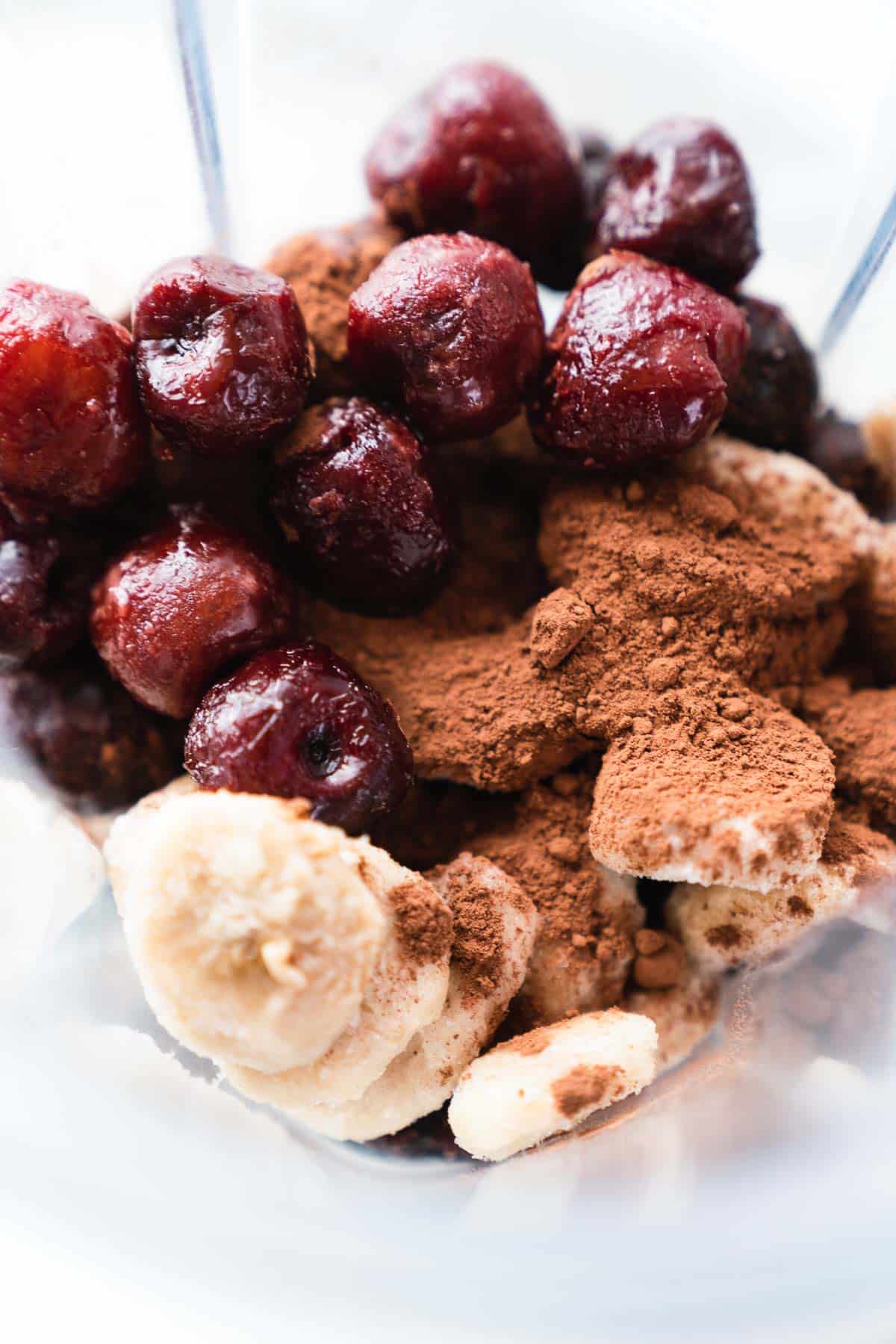 Frozen cherries, bananas and cocoa powder in a clear blender cup.