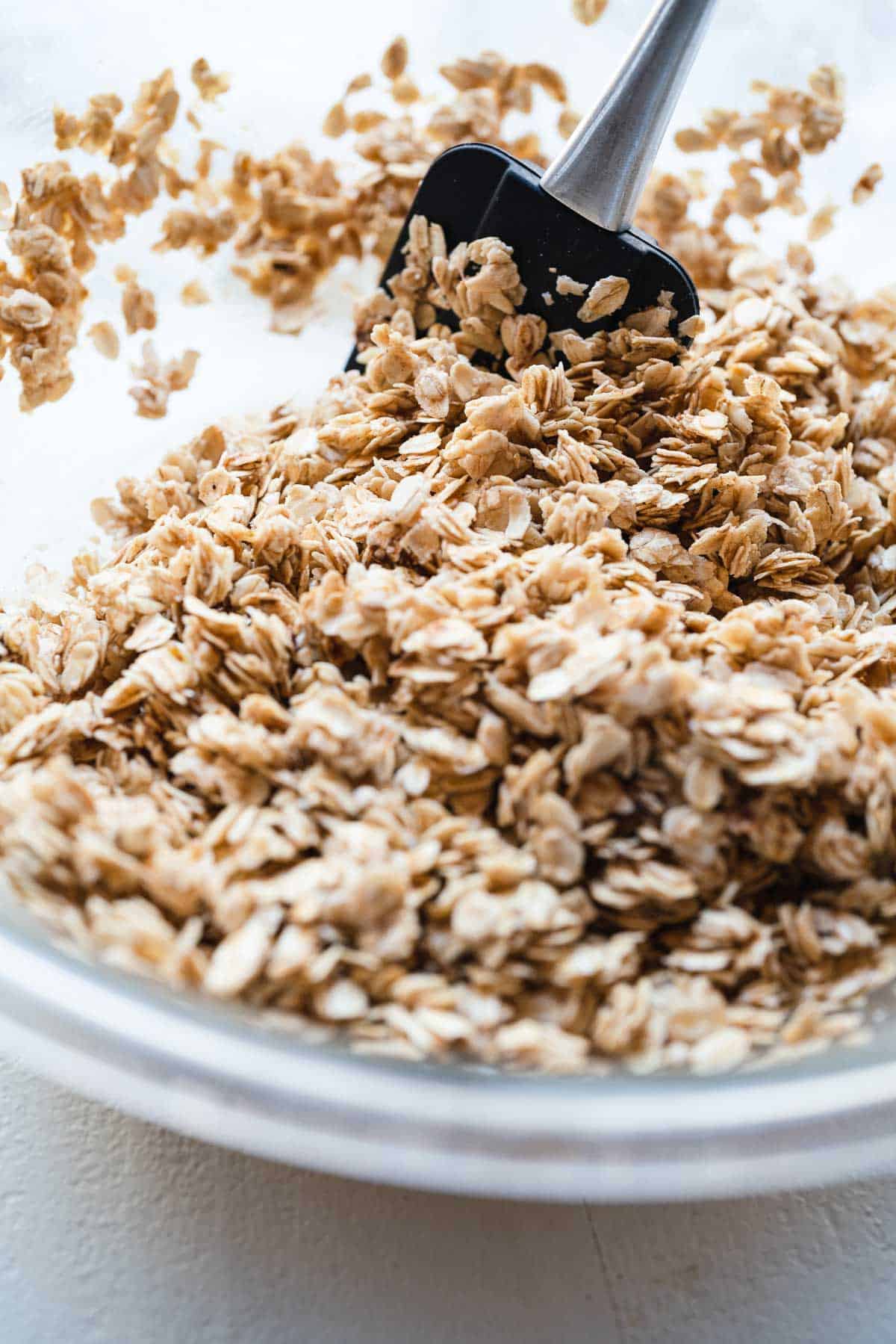 Oats being stirred with a black spatula in a clear glass mixing bowl.