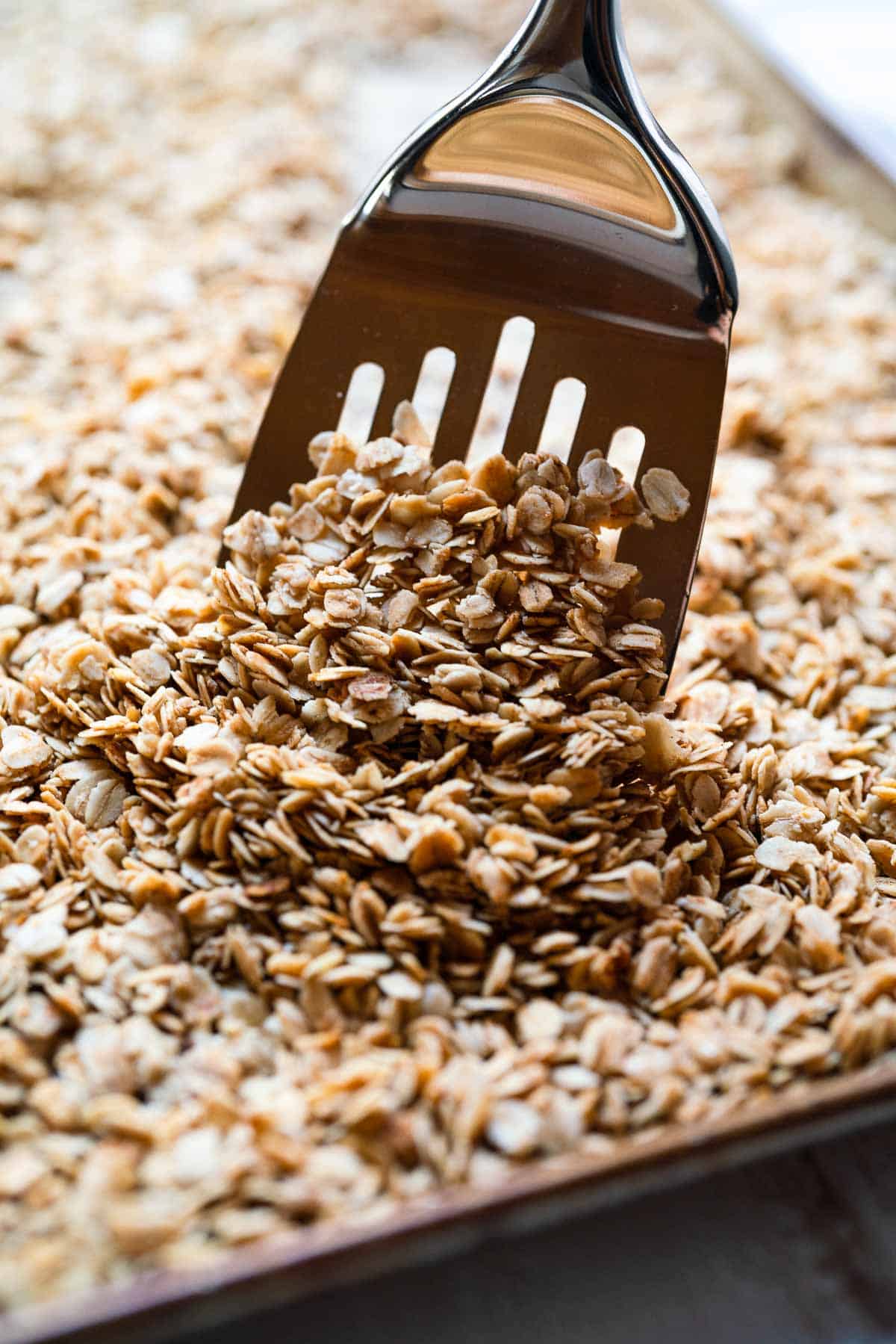 A gold spatula stirring toasted oat granola on a sheet tray.