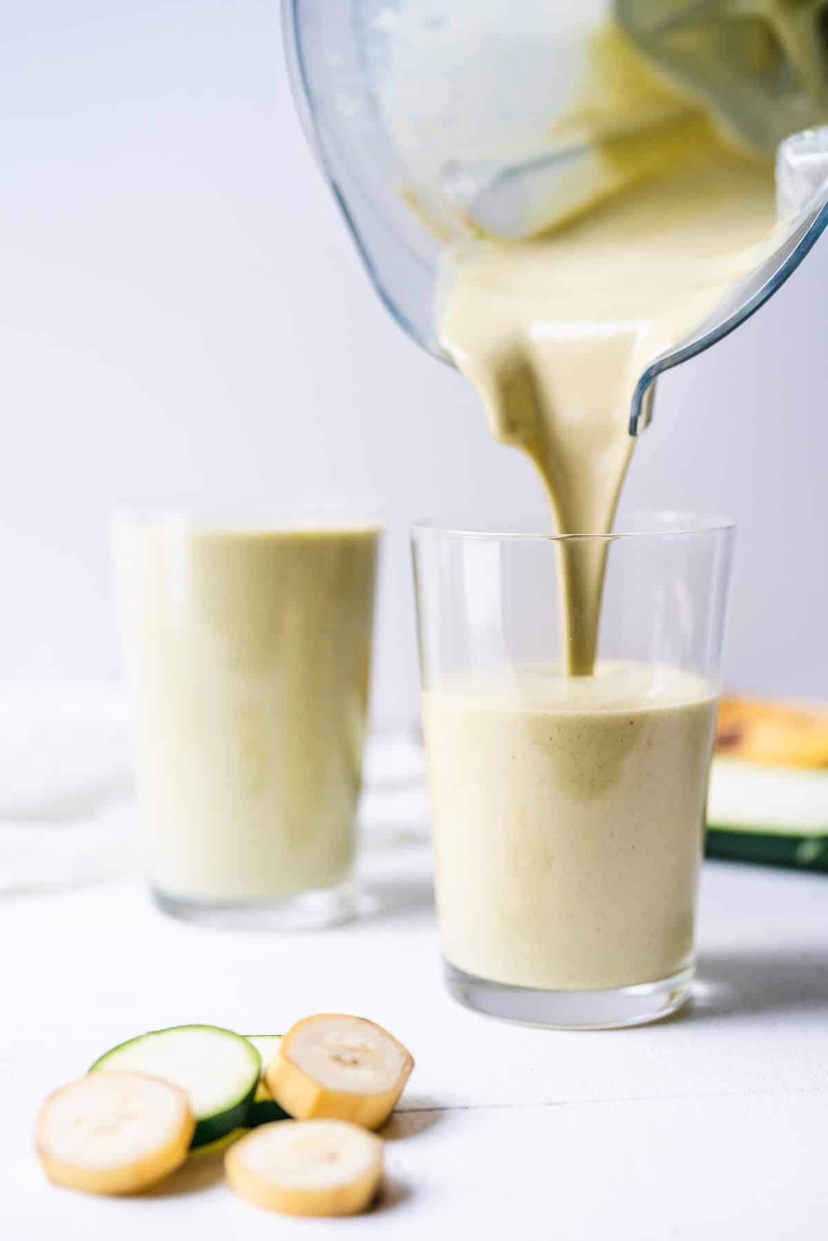 Two glasses being filled with a green smoothie with sliced bananas in the front.
